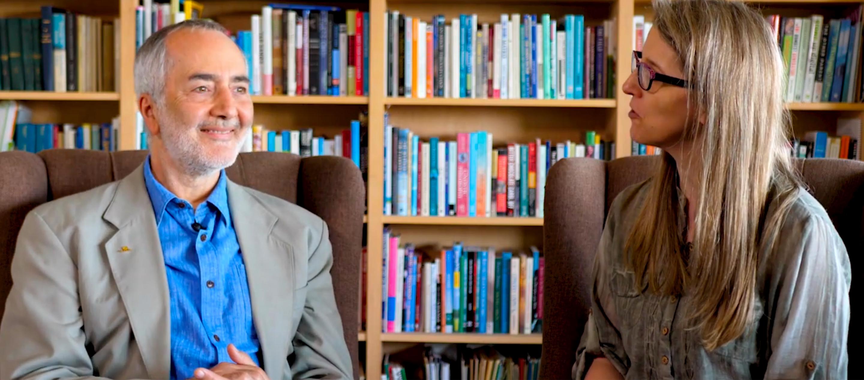 Raffi is shown sitting in an armchair in front of a shelf full of books during an interview 