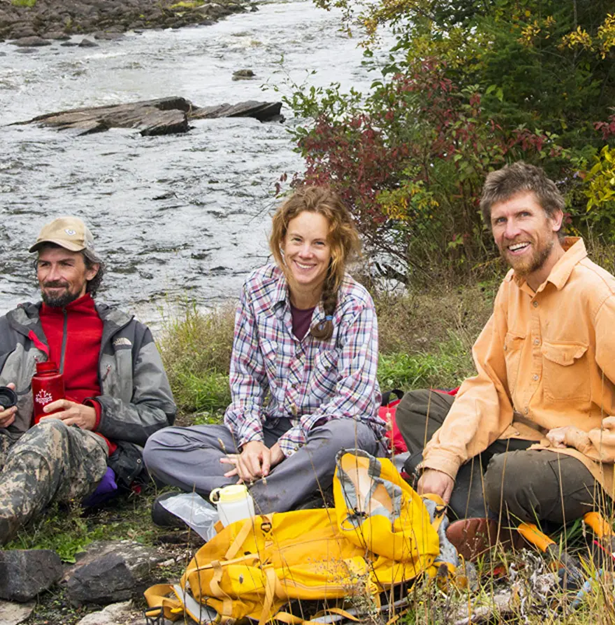 three people sitting beside a river all smiling at the camera
