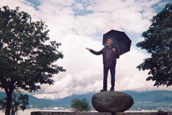 A man is seen standing under an umbrella on a large boulder next to the seawall. He is holding out an outstretched arm as if to check for rain. The sky above him is cloudy.