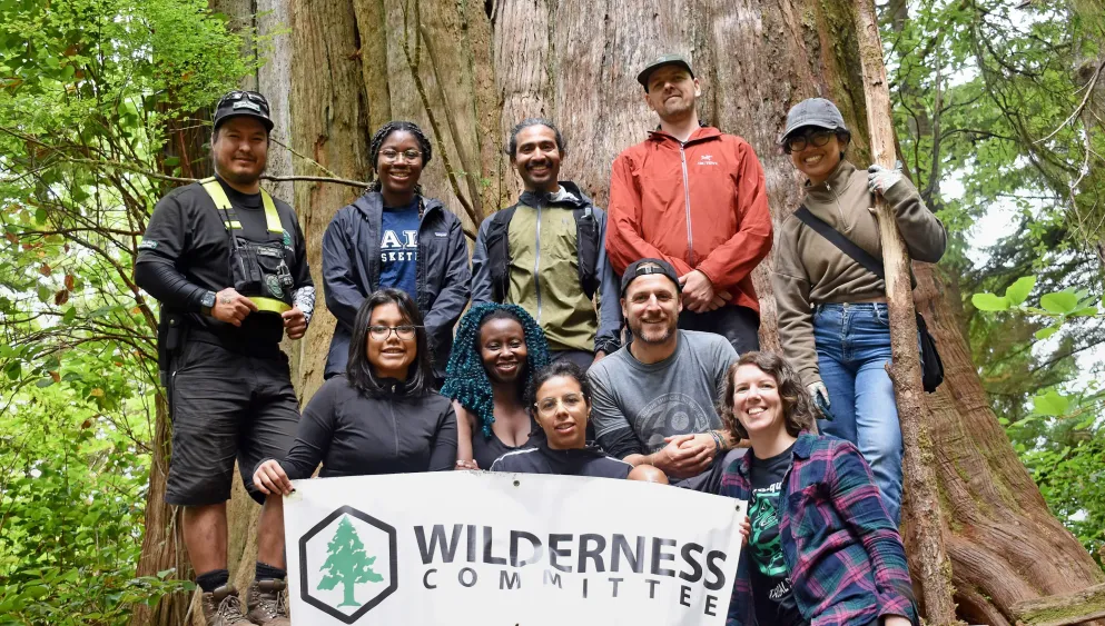 Wilderness Committee Group standing in front of a large tree with a sign featuring the company logo