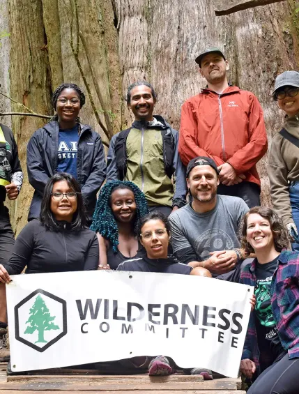 Wilderness Committee Group standing in front of a large tree with a sign featuring the company logo